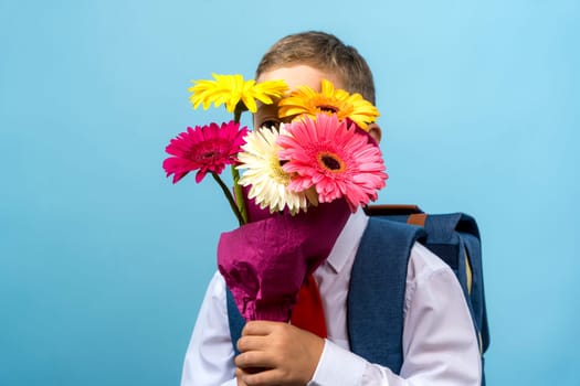 a funny first grader in a white shirt with a backpack holds a bouquet of flowers in his hands and cunningly looks out from behind the bouquet looking at the camera. Cute Caucasian boy goes to school. Schoolboy. September 1st. for the first time in the first grade. blue background