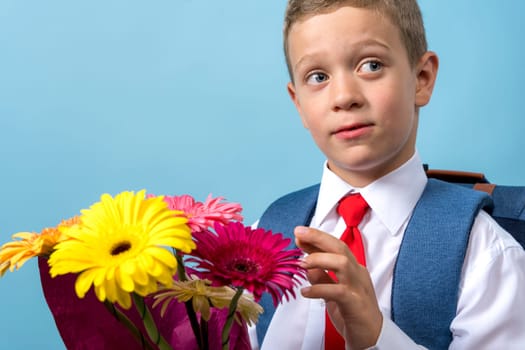 a first grader in a white shirt with a backpack holds a bouquet of flowers in his hands. Cute Caucasian boy goes to school with a bouquet of flowers and a backpack. Schoolboy. September 1, school education. for the first time in the first grade and a bouquet of flowers to the teacher, blue background