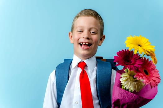 a happy funny first grader in a white shirt with a backpack holds a bouquet of flowers in his hands and laughs. Cute Caucasian boy goes to school. Schoolboy. September 1, for the first time in the first grade. blue background
