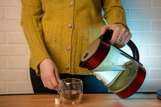 Woman poring hot boiled water from electric kettle into a clear glass cup on table. The process of brewing tea or tea ceremony in a warm soft light. Movement of boiling water pouring into a cup