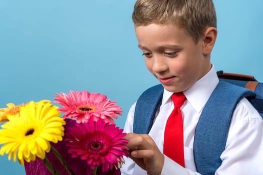 a happy funny first grader in a white shirt with a red tie examines and touches a bouquet of flowers in his hands. Cute Caucasian boy goes to school. Schoolboy. September 1, for the first time in the first grade. blue background