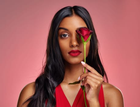 Face, beauty and portrait of a woman with a rose on a studio background for valentines day. Makeup, model and serious young Indian girl with a flower in hand for romance or love on pink backdrop.