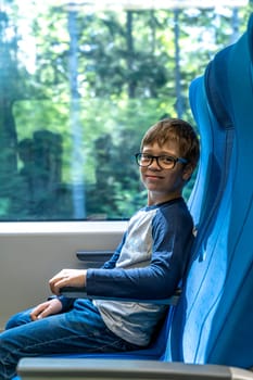 a cute Caucasian school-age boy with glasses rides a commuter train, sitting at the window. the boy smiles and looks at the camera for a fun journey. Have fun while traveling on public transport. Holidays, trip to school