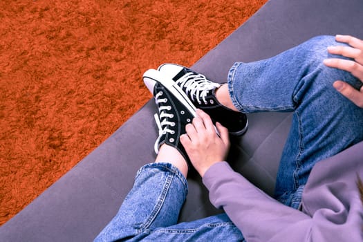 A happy relaxed girl is resting sitting on a comfortable sofa in the living room of the house. A pretty girl in jeans, casual sneakers and a hoodie is sitting at home on the couch.