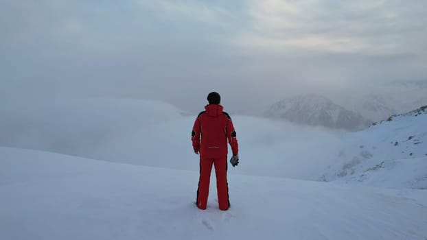 A climber in a red suit stands at the top of the mountain. Looks at the heavenly ocean of white clouds. There is snow all around, blue sky, dawn and calm. An epic moment of life. There are tours