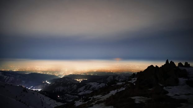 View of the night city from the snowy mountains. The glow from the city rises to the clouds. Bright lights are visible. In places, even the stars are visible. Steep cliffs, big rocks and snow. Almaty