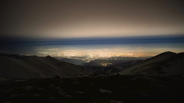 View of the night city from the snowy mountains. The glow from the city rises to the clouds. Bright lights are visible. In places, even the stars are visible. Steep cliffs, big rocks and snow. Almaty