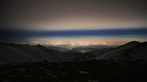 View of the night city from the snowy mountains. The glow from the city rises to the clouds. Bright lights are visible. In places, even the stars are visible. Steep cliffs, big rocks and snow. Almaty