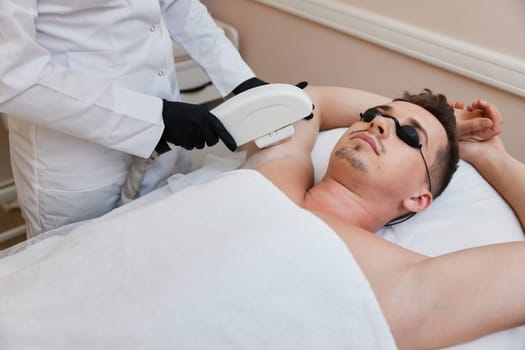 Young man receiving epilation in beauty center. Professional beautician doing laser hair removal procedure on male face.