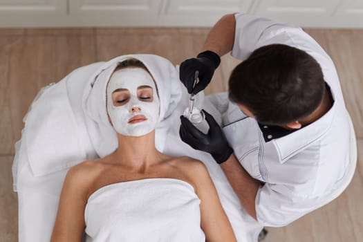 male beautician applies the mask to the face of beautiful woman in the spa salon. facial skin care concept.