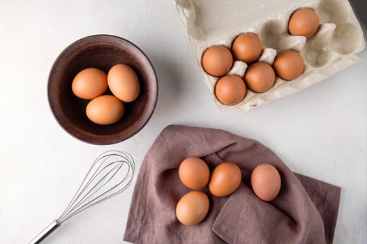 View from above brown chicken eggs in a cardboard packing container, and in a bowl on a table with a whisk to beat the eggs.