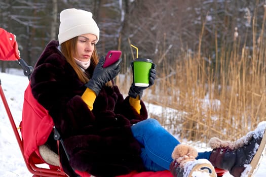 an adult female human being. Pretty woman with smart phone and hot coffee tea sits on sled in winter forest.