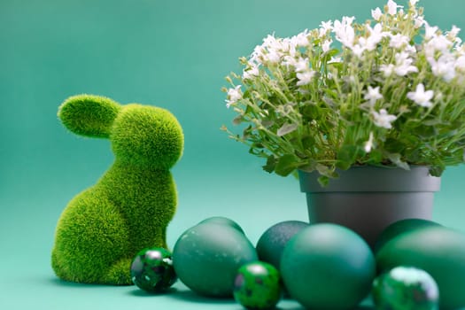 Closeup Easter green bunny, rabbit with blue colored chicken quail eggs on blue background. White spring flowers in pot. Pascha, Resurrection Sunday, Christian festival, cultural monochrome holiday.