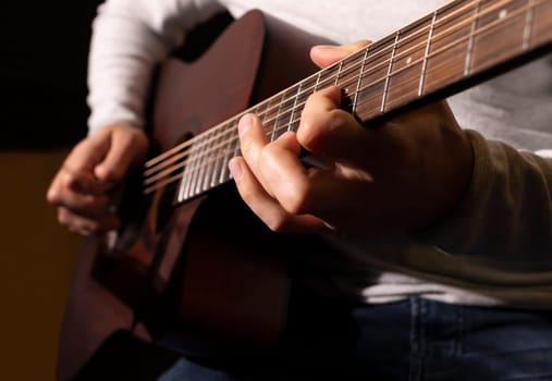 Closeup white man plays guitar, holding musical instrument in hands, sitting on chair in studio. Darken photo. Male wears casual cloth. Hobby, leisure of creative person, artist. Horizontal plane.