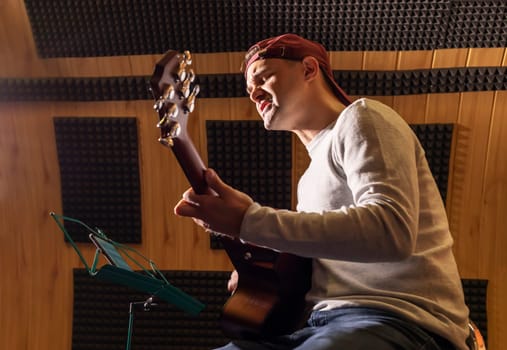 Caucasian man sings song, playing guitar, holding musical instrument in hands, sitting in chair in studio. Darken photo. Male wears casual cloth. Hobby, leisure of creative person, artist.Horizontal