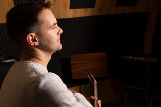 Caucasian man plays guitar, holding musical instrument in hands, sitting in chair in studio. Darken photo. Male wears casual cloth. Hobby, leisure of creative person, artist. Copy space. Horizontal