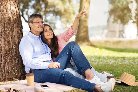 Romantic Middle Aged Couple Having Picnic In Summer Park, Happy Mature Man And Woman Sitting On Plaid Under Tree, Loving Spouses Enjoying Takeaway Coffee, Looking And Pointing Away, Copy Space