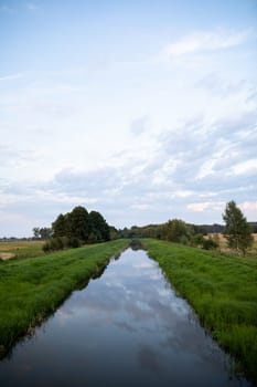 The river with a very slow current flows straight amidst green grasses and deciduous trees. The overcast sky is reflected in the calm river.