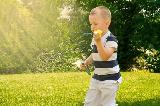 A handsome caucasoid boy 4 years old, in a striped T-shirt, plays outdoors in a green park in summer and blows soap bubbles in the sun. Childhood concept.