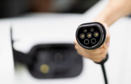 Progressive new alternative clean energy vehicle and electric charging device concept. Focus closeup hand holding and pointing EV charger plug at camear from electric charging station at camera