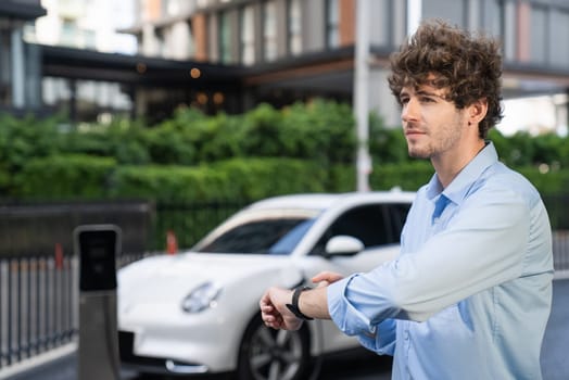 Businessman with smartwatch at modern charging station for electric vehicle with background of residential buildings as concept for progressive lifestyle of using eco-friendly as alternative energy.