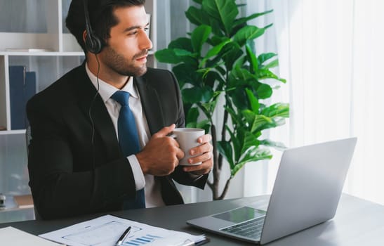 Manager of call center operator office sitting on his desk with his coffee while working on laptop. Office worker wearing headset and black suit working on customer support or telemarketing. fervent