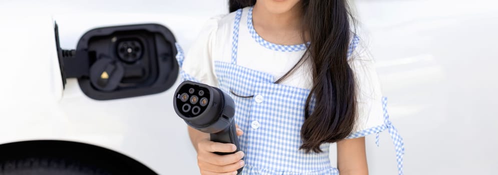 A playful girl holding and pointing an EV plug, a home charging station providing a sustainable power source for electric vehicles. Concept of progressive new generation with ecological awareness.