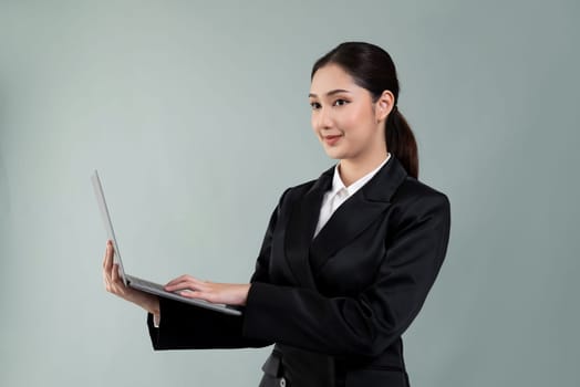 Confident young businesswoman stands on isolated background, working on laptop posing in formal black suit. Office lady or manager with smart and professional appearance. Enthusiastic