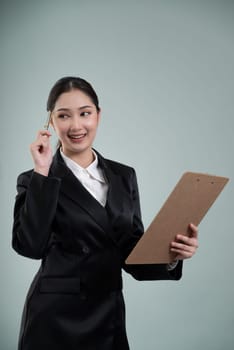 Confident young businesswoman stands on isolated background, holding clipboard and posing in formal black suit. Office lady or manager with smart and professional appearance. Enthusiastic