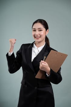 Confident young asian businesswoman in formal suit holding clipboard with hand gesture indicating promotion or advertising with surprised facial expression on isolated background. Enthusiastic