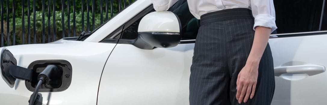 Closeup progressive suit-clad businesswoman with her electric vehicle recharge her car on public charging station in modern city with power cable plug and renewable energy-powered electric vehicle.