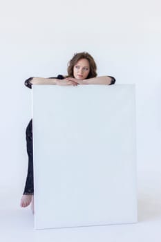 Young Woman Leans On White Blank Large Stretched Canvas for Painting. Beautiful Female Artist Poses With Empty Boards On White Background Indoor. Mockup. Vertical Plane. High quality photo