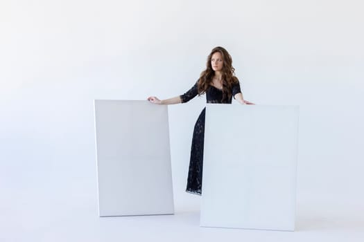 Young Woman Stands Between White Blank Large Stretched Canvases for Painting. Beautiful Female Artist Poses With Empty Boards On White Background Indoor. Mockup. Horizontal Plane. Copy Space.