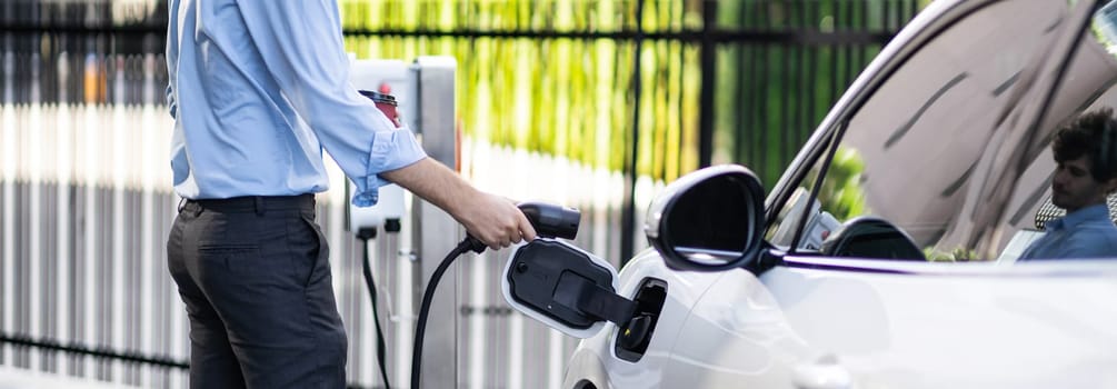 Closeup progressive man holding EV charger plug from public charging station for electric vehicle with background of residential building as concept eco-friendly sustainability energy car concept.