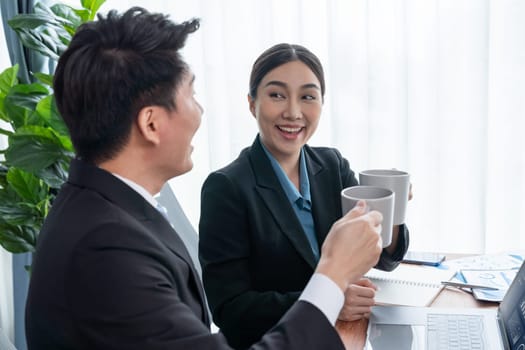 Two Asian office workers taking coffee break together in workplace. Coworkers smiling and socializing while holding cup of coffee adding friendly working environment in corporate workspace. Jubilant