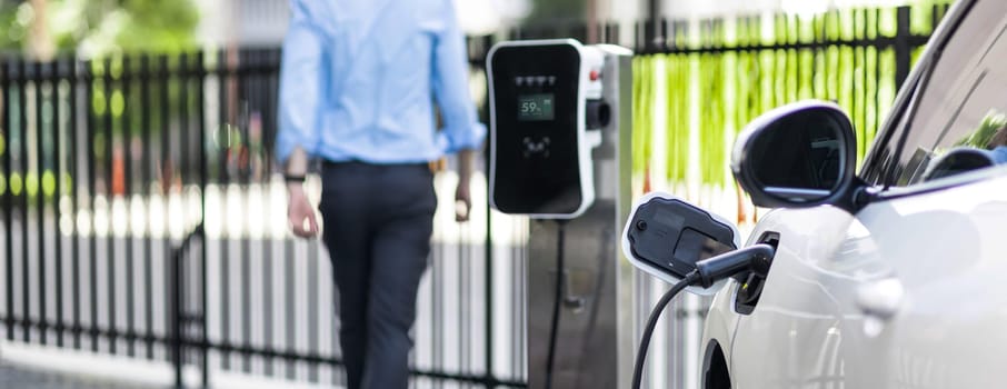 Focus closeup electric vehicle recharge battery at public charging station in the city area with blur businessman in background. EV car attached with electric charger for eco-friendly idea.