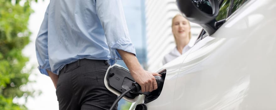 Closeup focus hand insert EV charger to electric vehicle at public charging point in car park with blur business people in backdrop, eco-friendly lifestyle by rechargeable car for progressive concept.