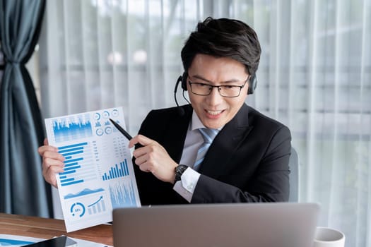 Confident asian telesales businessman work in modern office wearing headphone, presenting BI dashboard analysis to clients and customers in online meeting. Success modern telesales business . Jubilant