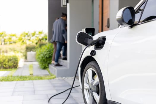 Focus closeup electric vehicle recharging battery from home electric charging station with blurred family in background. Renewable clean energy car for progressive eco awareness lifestyle concept.