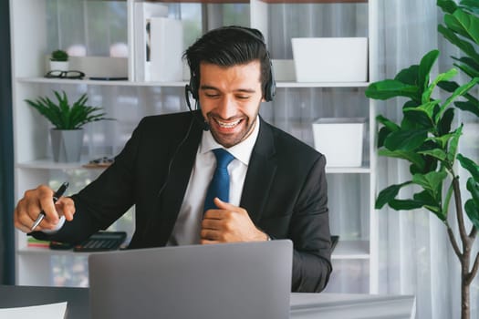 Excited and happy businessman dressed in black formal suit, raise his arm in celebratory gesture at office desk. Successfully call center or telesales operator celebrate in workplace. Fervent