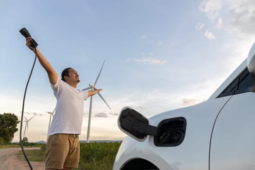 Progressive man spread arm with electric car recharge energy from charging station on green field powered by wind turbine concept of future sustainable energy.