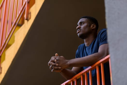 Portrait African American Young Sportive Man Standing On Balcony, on Stairs Looking Away Outdoor. Male Puts Hands On Orange Railing, Yellow Building. Horizontal Plane. Copy Space For Text.