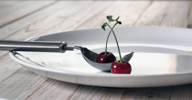 Red cherries in a white plate with a spoon for laying out ice cream on a white wooden table.Food background
