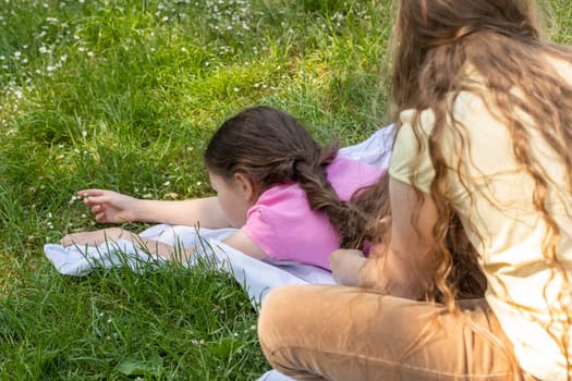 View From Back Little Girl With Long Hair braids Sister's Hair Sitting On Grass in Meadow At Sunny Day. Siblings Love, Carefree Time, True Friendship, Family Lifestyle. School Break. Horizontal Plane