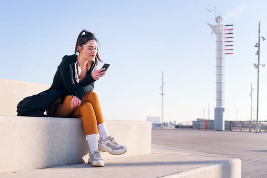 young sportswoman sitting outdoors in the city using a mobile phone, concept of technology of communication and urban lifestyle, copy space for text