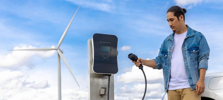 Progressive man with his electric car, EV car recharging energy from charging station on green field with wind turbine as concept of future sustainable energy. Electric vehicle with energy generator.