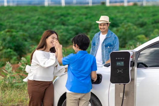 Concept of progressive happy family enjoying their time at wind farm with electric vehicle. Electric vehicle driven by clean renewable energy from wind turbine generator for charging station.