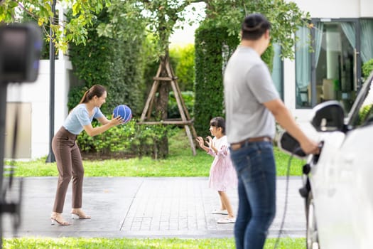 Progressive young happy family with electric vehicle and home backyard charging station. Green and clean energy from electric vehicles for healthy environment. Eco power from renewable source.