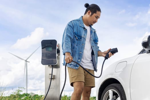 Progressive man with his electric car, EV car recharging energy from charging station on green field with wind turbine as concept of future sustainable energy. Electric vehicle with energy generator.