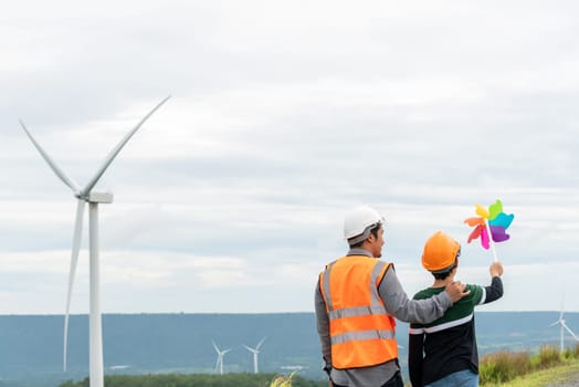 Engineer with his son holding windmill toy on a wind farm atop a hill or mountain. Progressive ideal for the future production of renewable, sustainable energy. Energy generated from wind turbine.